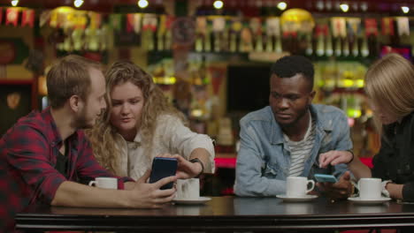 Portrait-of-cheerful-young-friends-looking-at-smart-phone-while-sitting-in-cafe.-Mixed-race-people-sitting-at-a-table-in-restaurant-using-mobile-phone